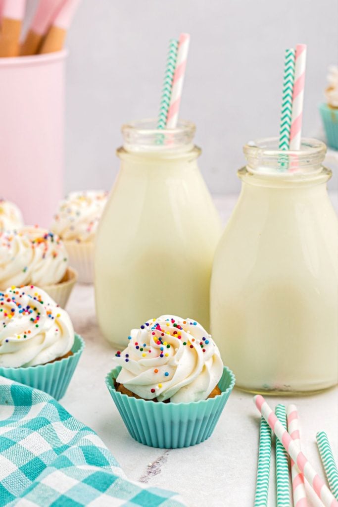 Frosted cupcakes in front of two bottles of milk with straws on a marble table. 