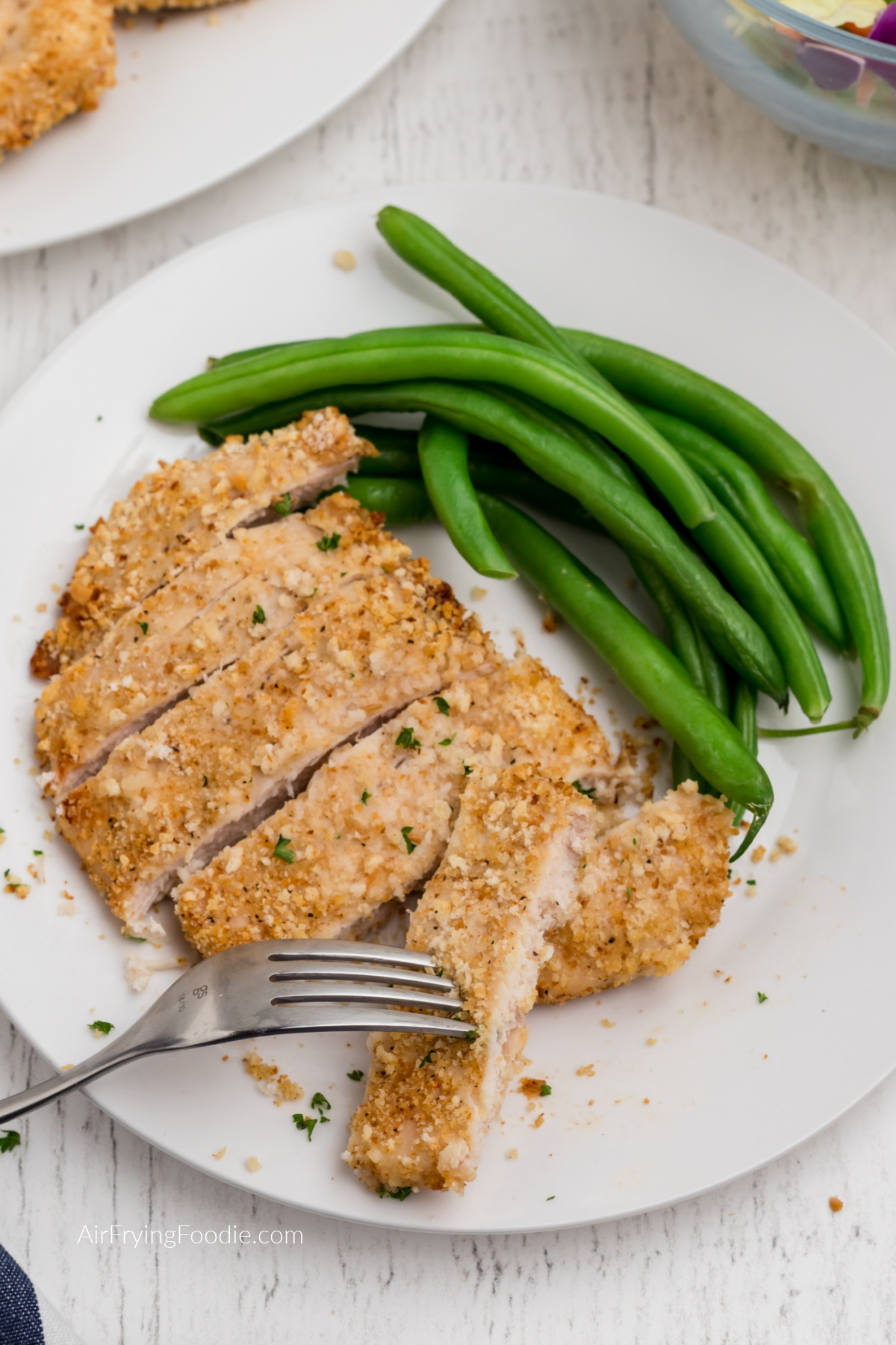 Sliced air fryer fried chicken with a fork taking a slice and a side of green beans on a white plate.