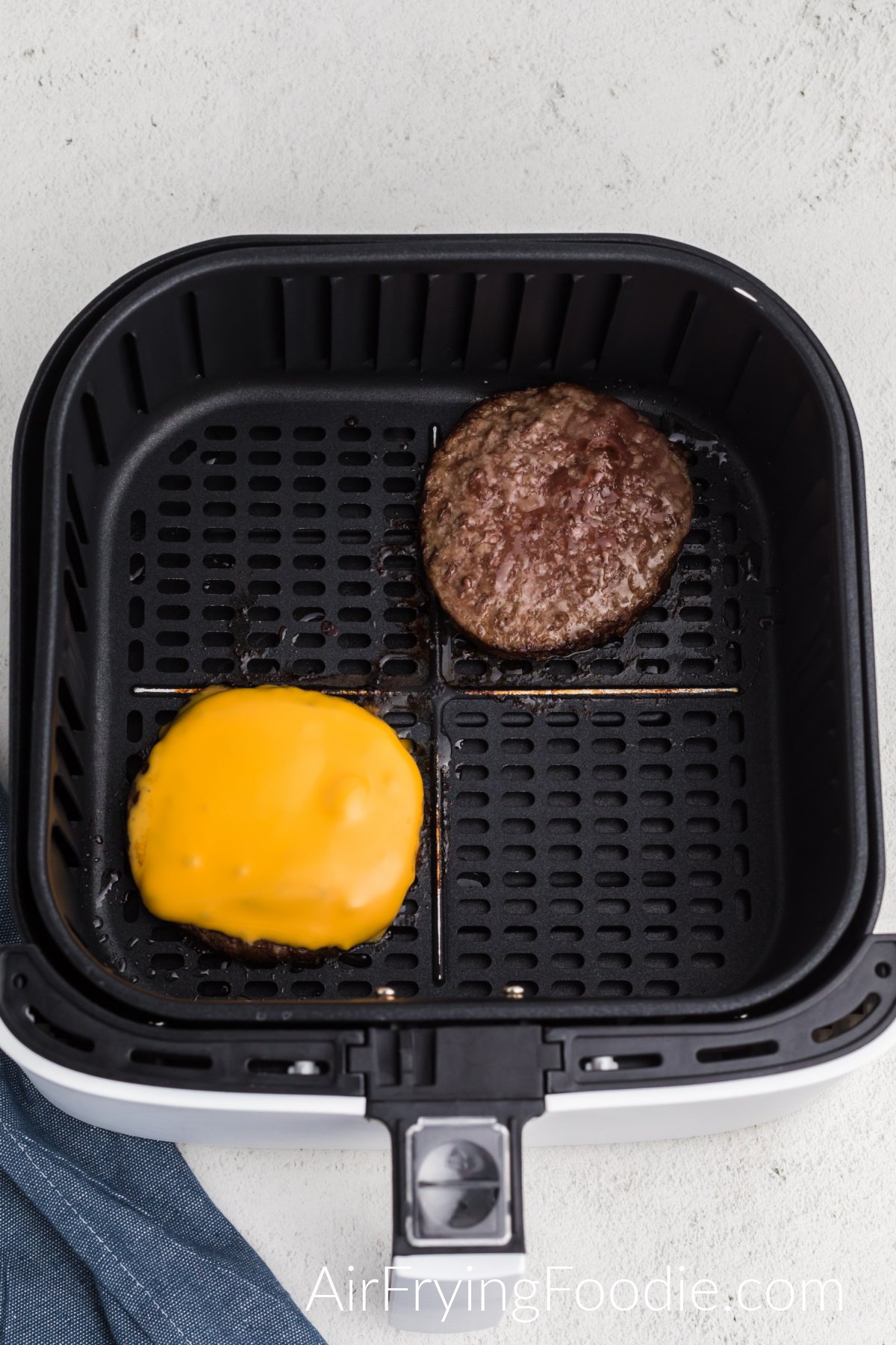 Cooked frozen burger in air fryer basket, one topped with sliced cheese.