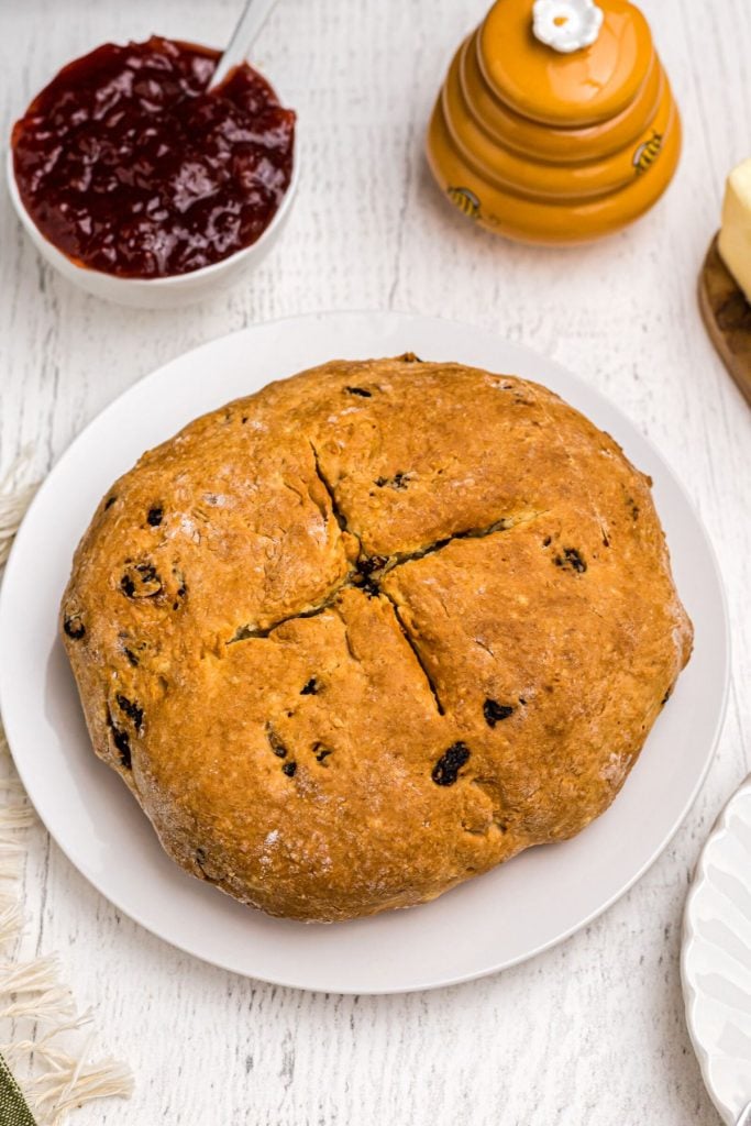 Golden brown round loaf of bread with raisins in it, on a white plate surrounded by butter, honey, and jam