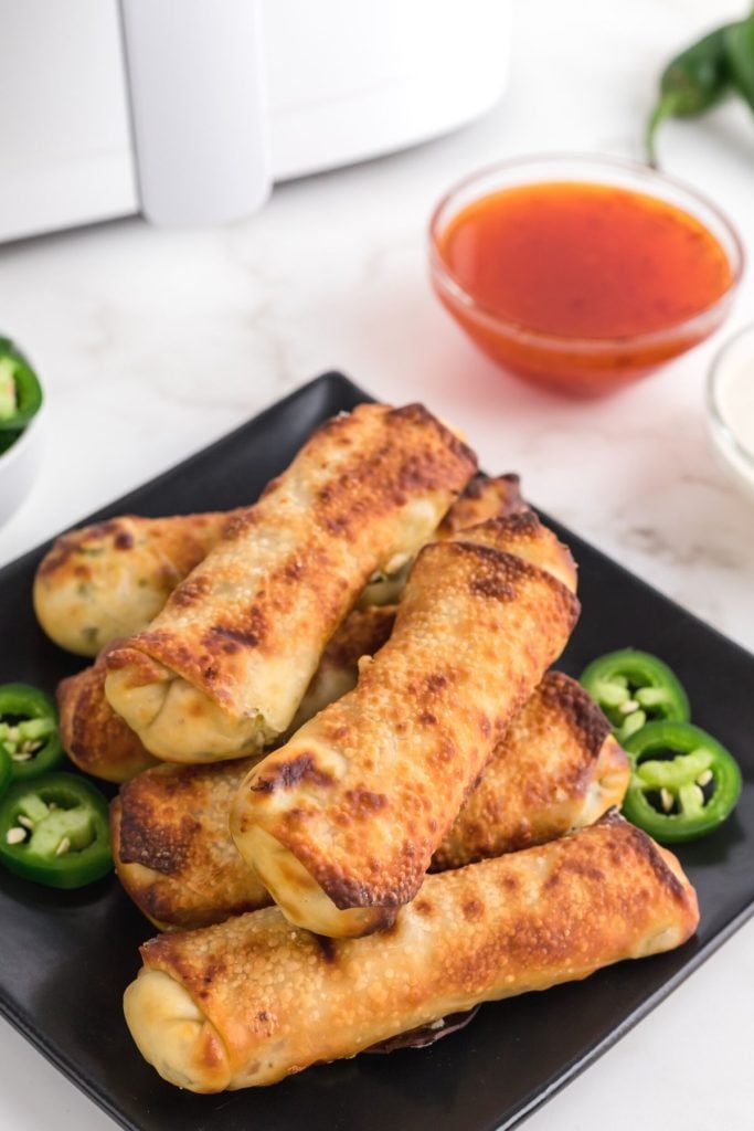 Golden crispy egg rolls on a black plate with jalapeno slices.