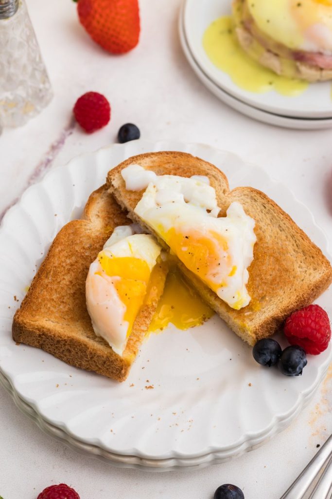 Poached egg on a piece of toast on a white plate with berries in front of a plate with eggs benedict. 