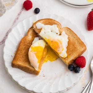 Poached egg on toast cut in half with yolk spilling out. Fresh fruit as a garnish on a white plate.