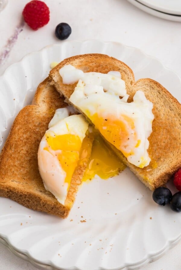 Poached egg on toast cut in half with yolk spilling out. Fresh fruit as a garnish on a white plate.