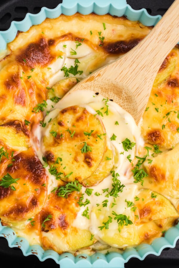 Cheesy scalloped potatoes being scooped from the dish in the basket of the air fryer.