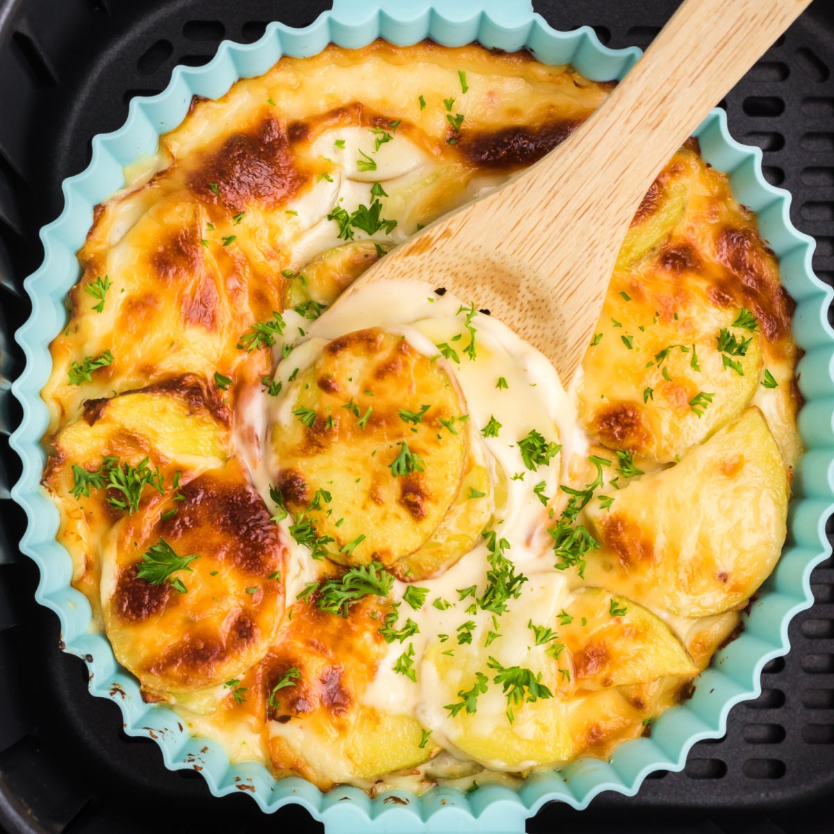Cheesy scalloped potatoes being scooped from the dish in the basket of the air fryer.