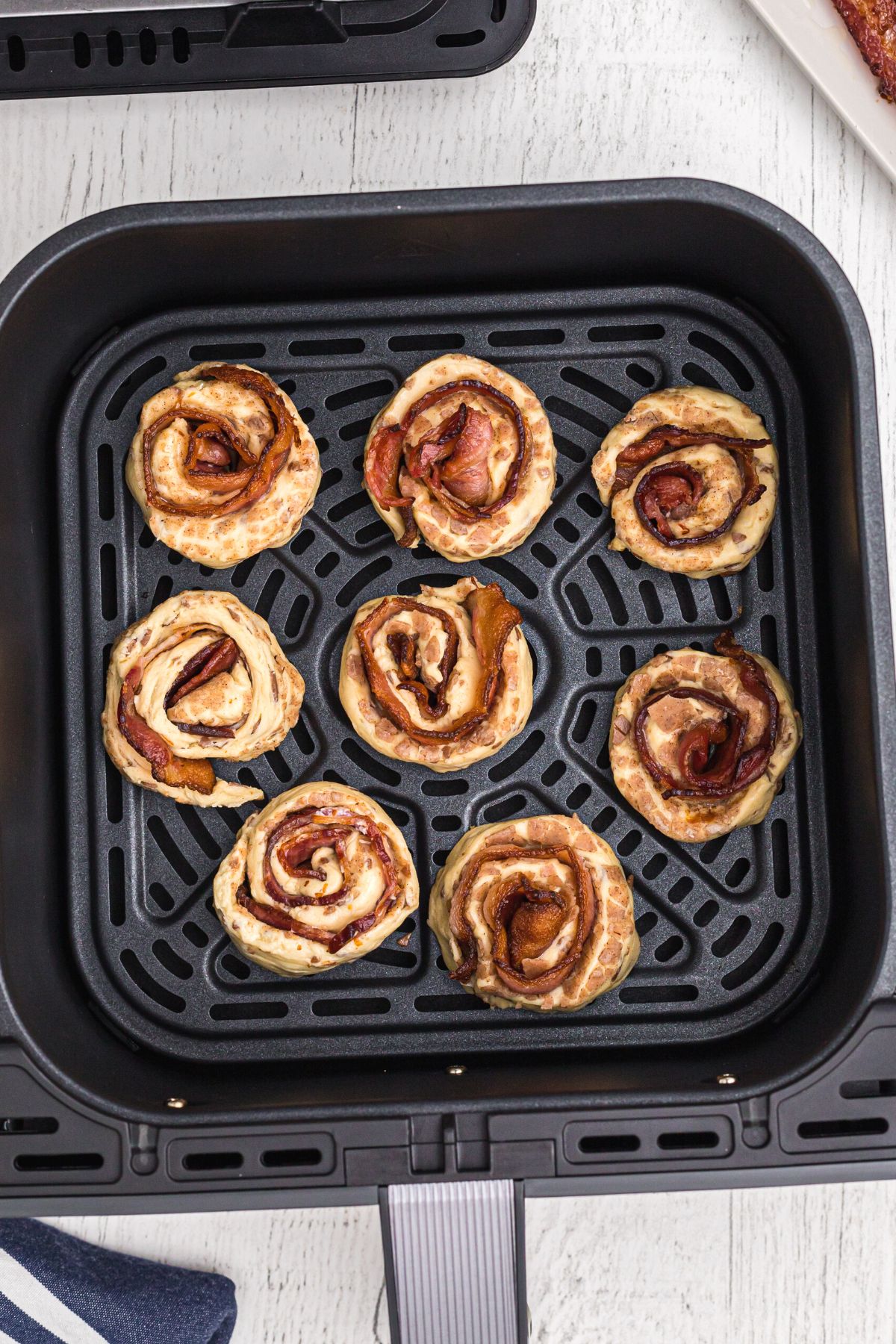 Uncooked cinnamon rolls wrapped around cooked bacon slices in the air fryer basket.