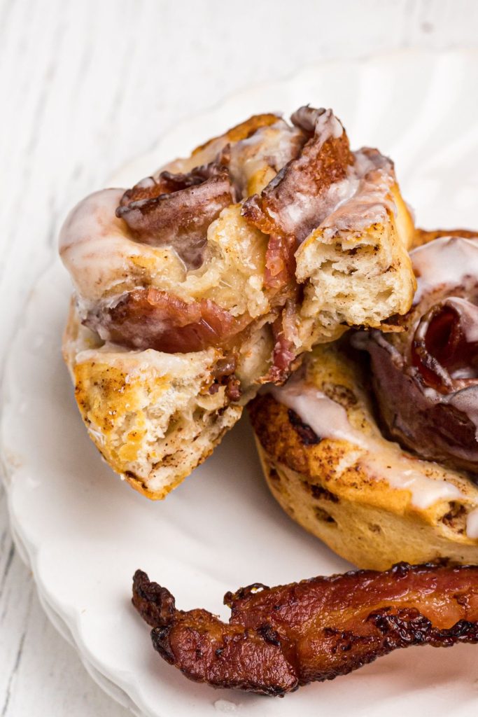 Golden cinnamon roll wrapped around a piece of cooked bacon, with a bite removed showing the inside of the roll. 
