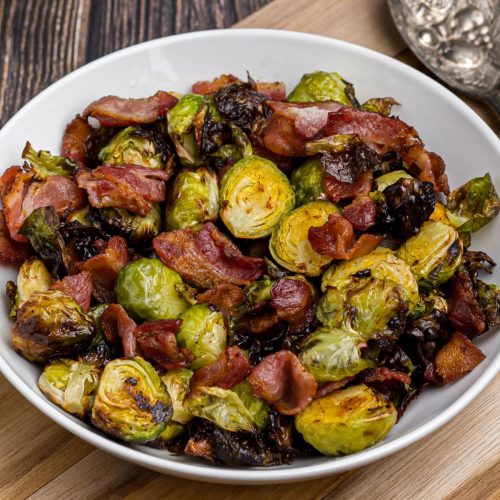 Crispy green brussels sprouts and bacon strips in a white bowl on a wooden cutting board.