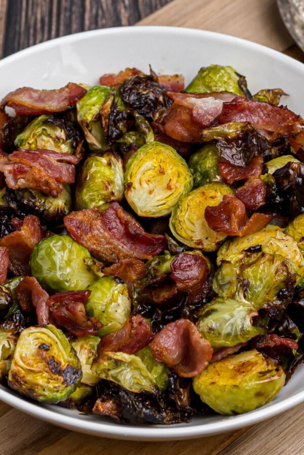 Crispy green brussels sprouts and bacon strips in a white bowl on a wooden cutting board.