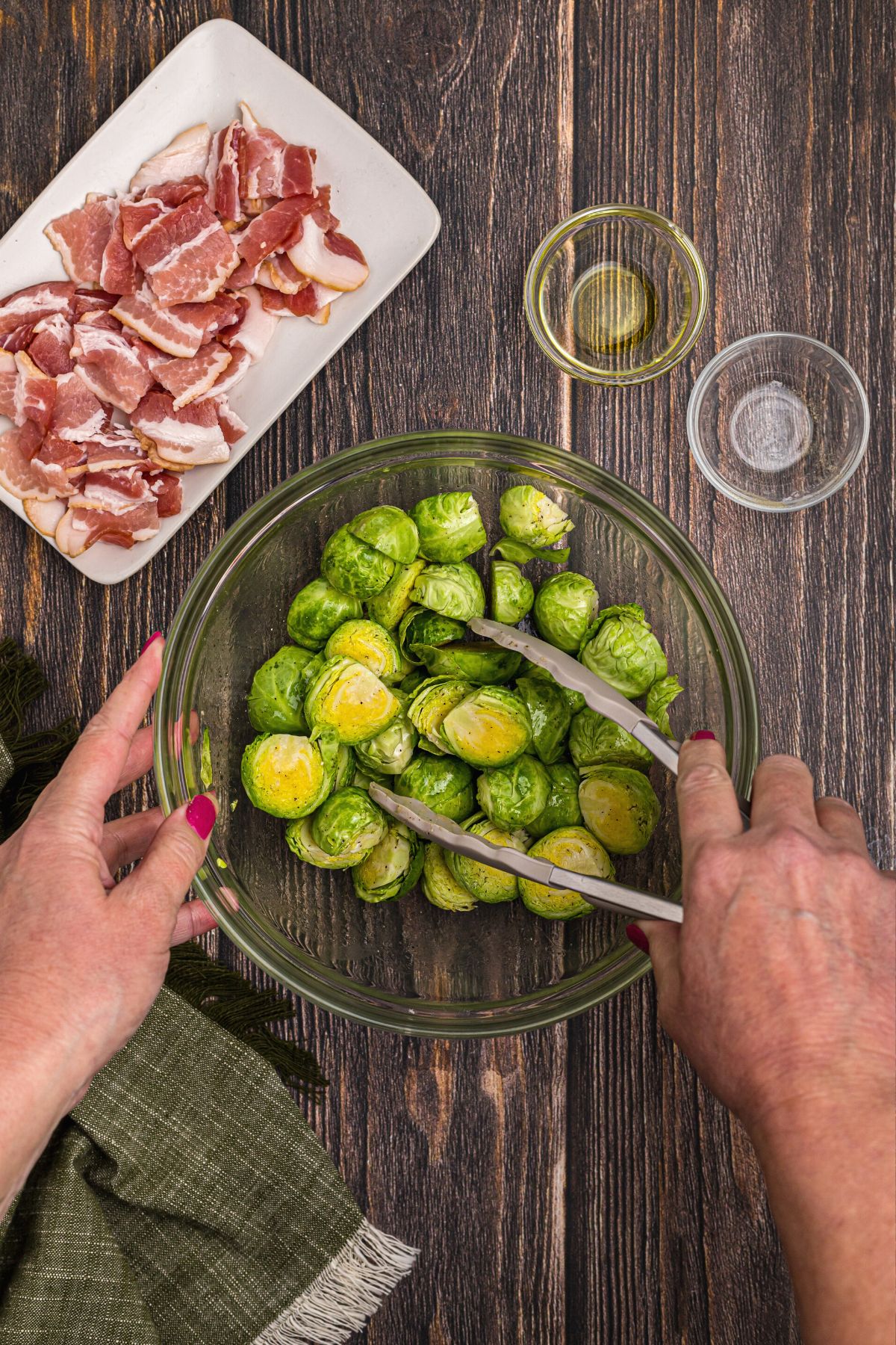 Cut green brussels sprouts being tossed with olive oil and seasonings in a clear mixing bowl on a wooden table.