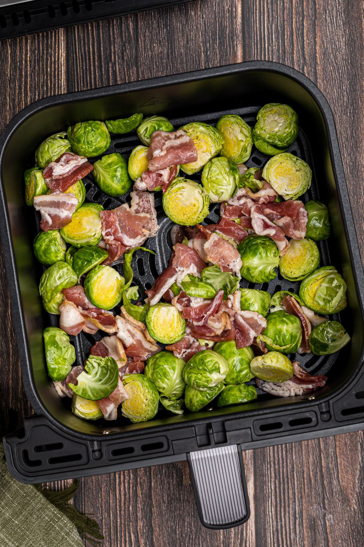 Uncooked seasoned brussels sprouts and bacon pieces in the air fryer basket.