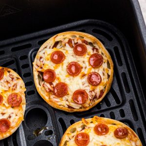 Golden crispy english muffin pizzas with melted cheese and cooked pepperonis in an air fryer basket.