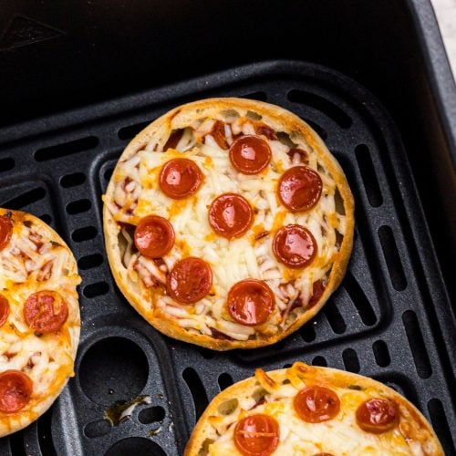 Golden crispy english muffin pizzas with melted cheese and cooked pepperonis in an air fryer basket.