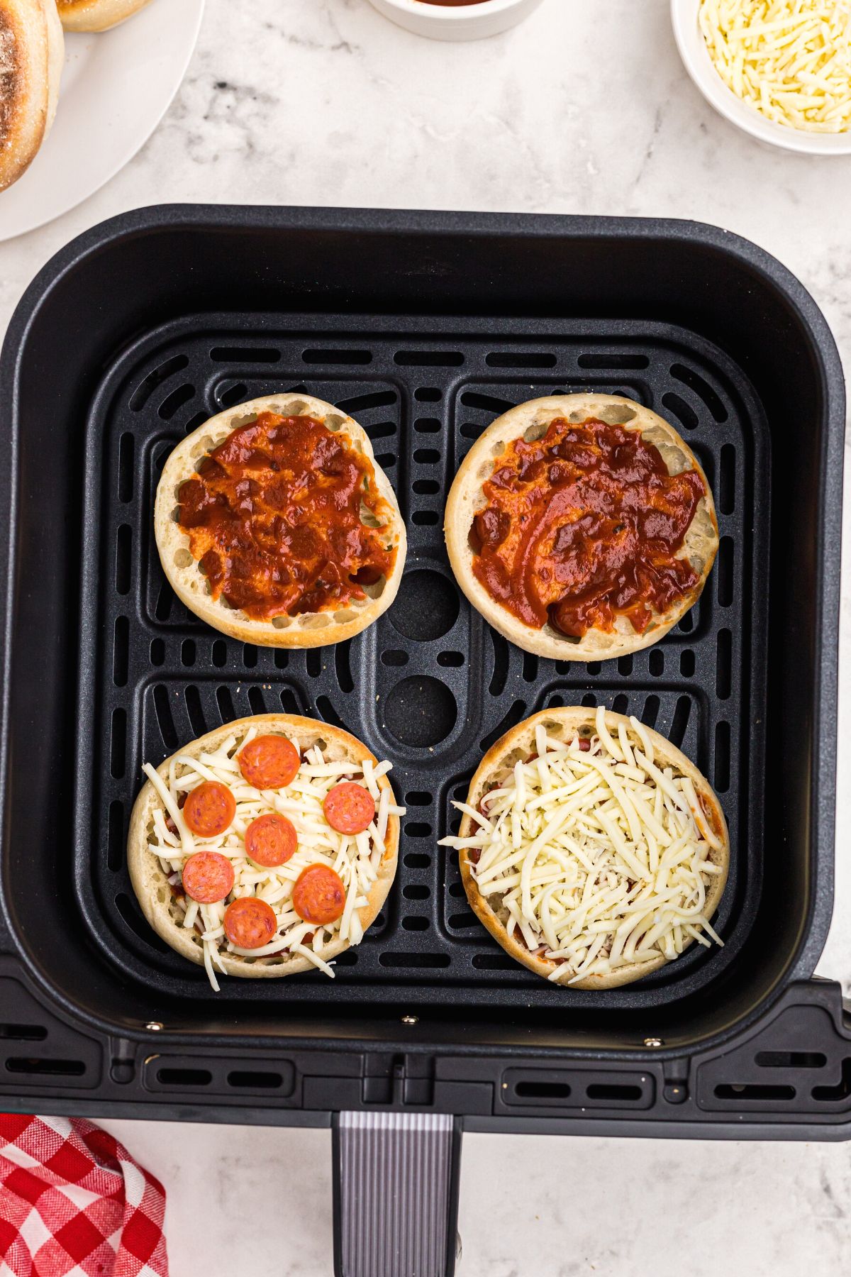 English muffin halves in the air fryer basket, topped with sauce, cheese, and pepperonis.