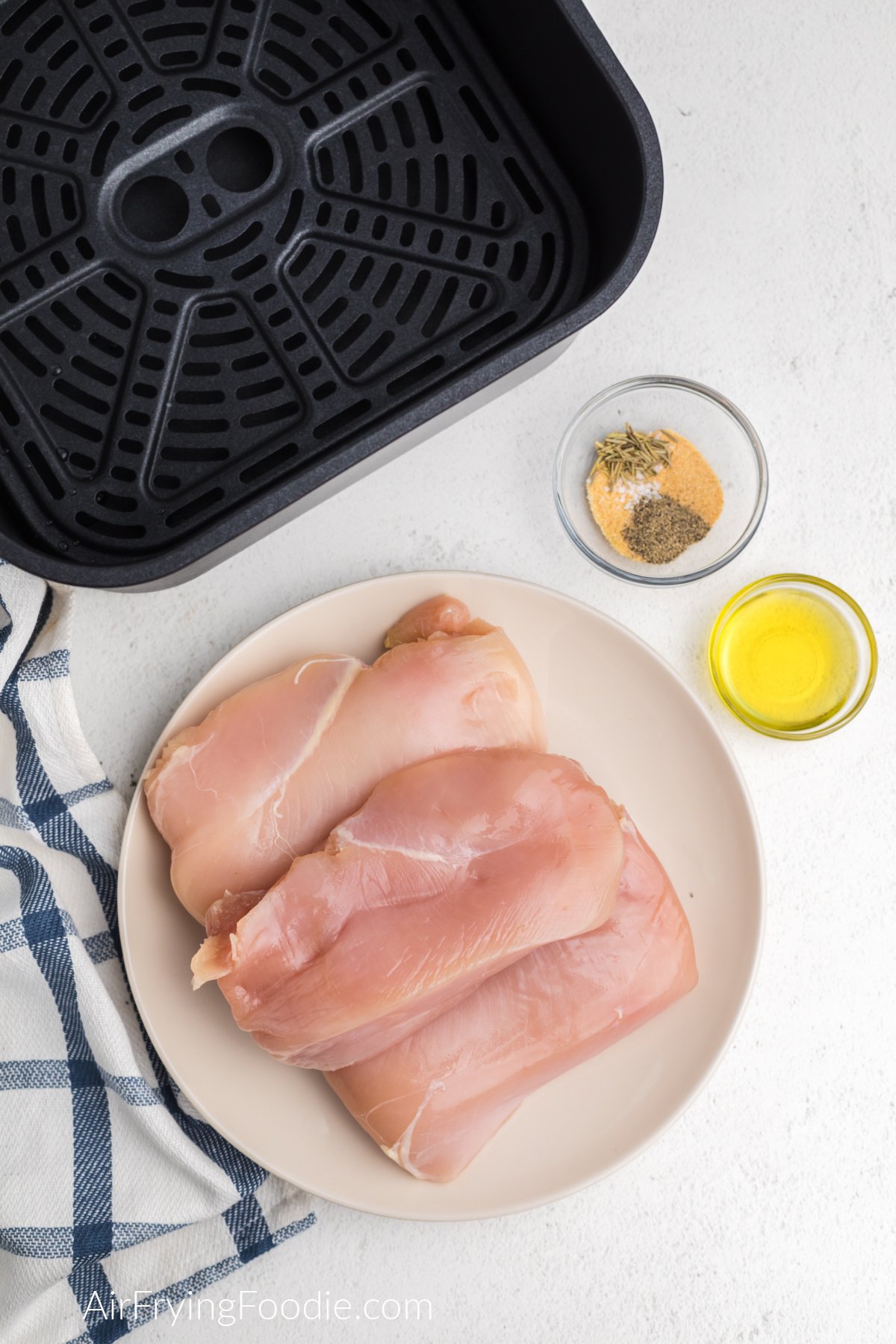 Boneless skinless chicken breasts on a white plate with a small bowl of olive oil and a bowl of seasonings. 