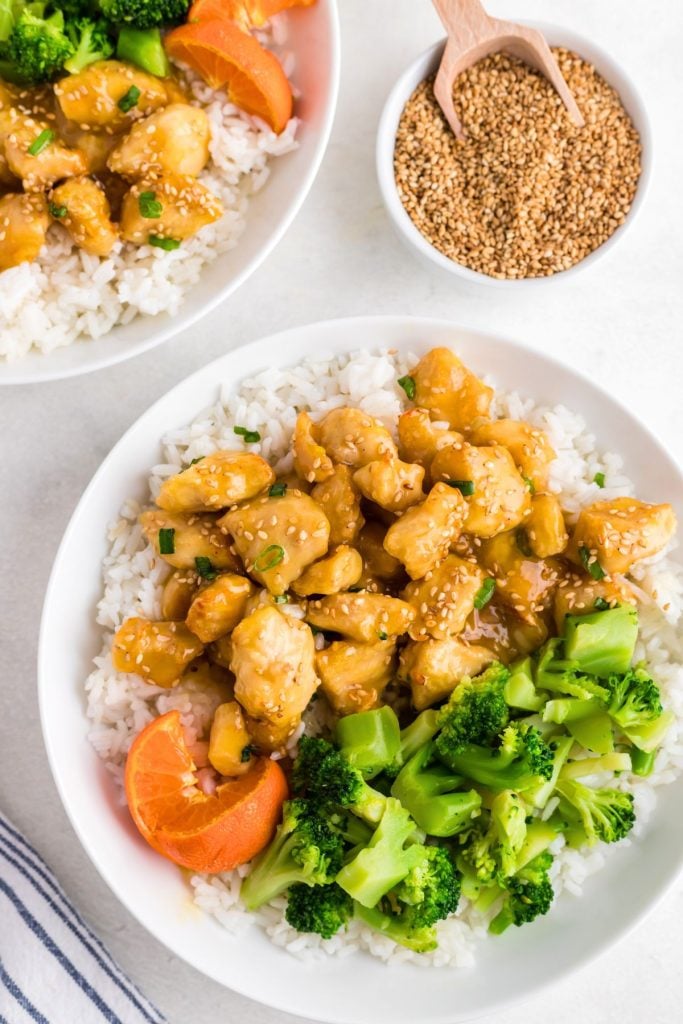 Golden orange chicken served on white rice with broccoli and an orange on a white plate. 