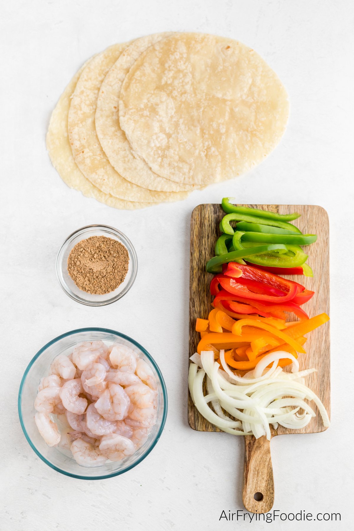 Shrimp, sliced onion and bell peppers on a board, seasoning, and tortillas on a table ready to be prepared.
