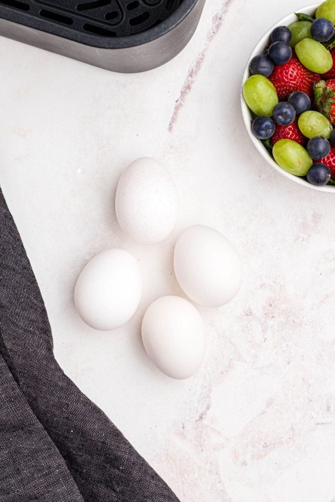 Four eggs on a marble table in front of an air fryer basket and a bowl of fruit. 
