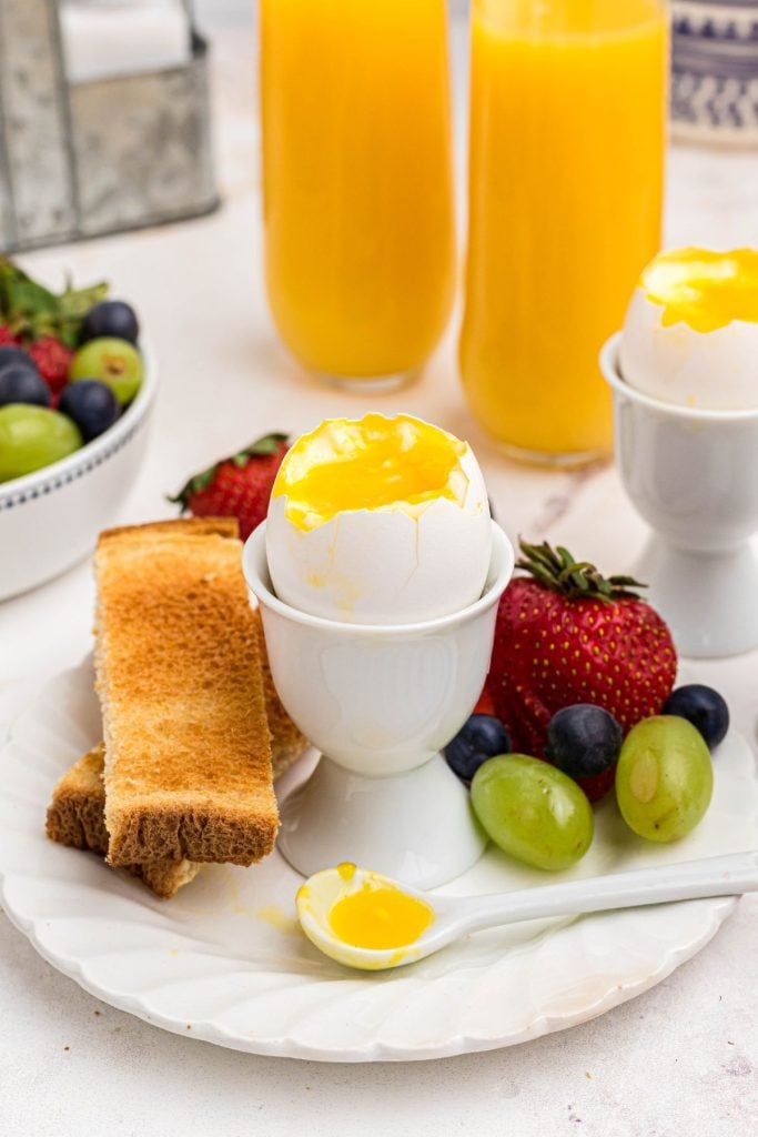 Slightly runny yolk inside of an egg shell in an egg holder with fruit and toast. 