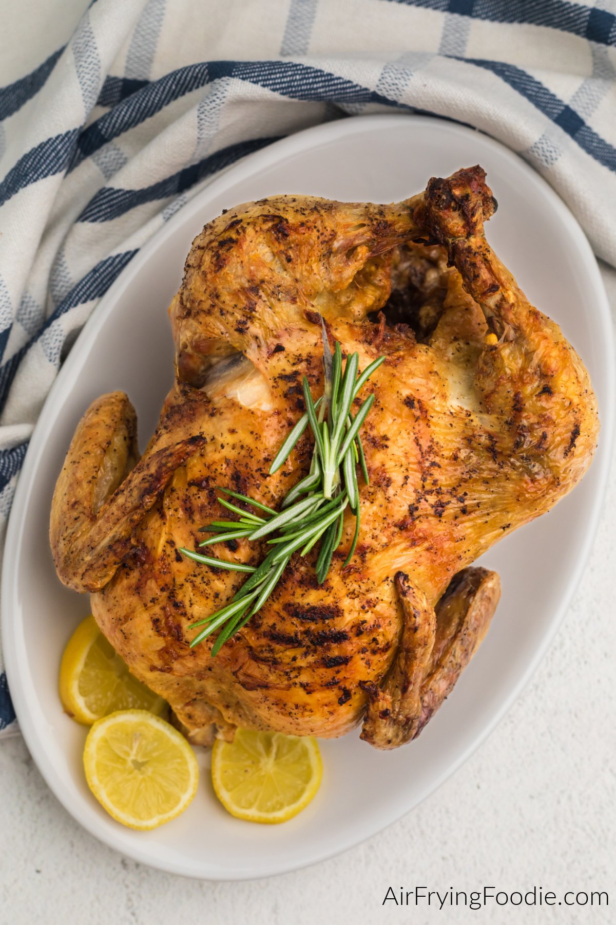 Air fried whole chicken on a white serving plate with sprigs of rosemary and lemon slices.