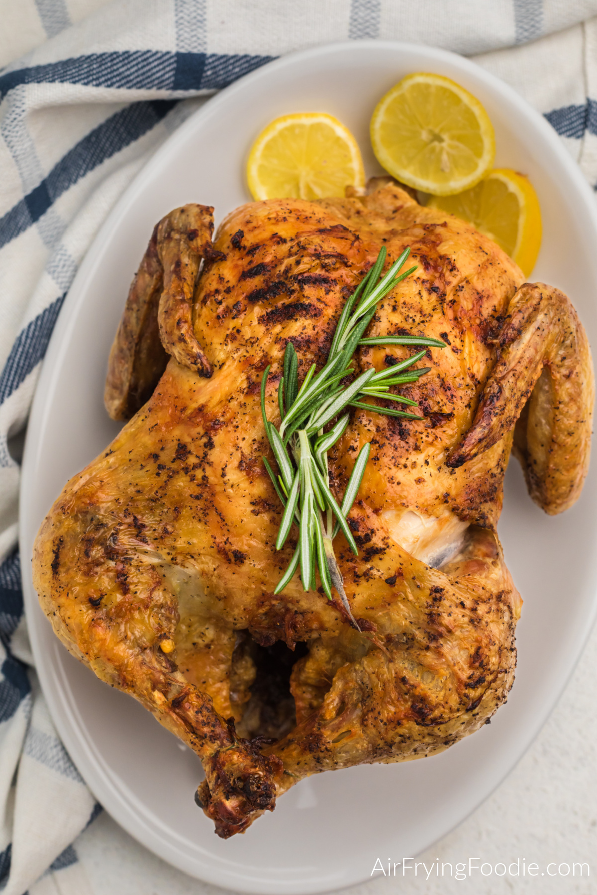 Air fried whole chicken on a serving plate with rosemary sprigs on the top and served with lemon slices. 
