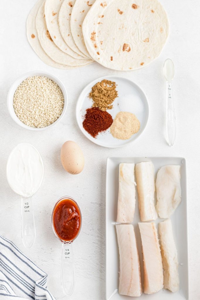 Uncooked fish, tortilla shells, egg, flour, breadcrumbs and seasonings measured out on a white table. 