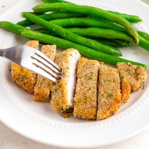 Golden breaded pork chop on a white plate with green beans as a side.