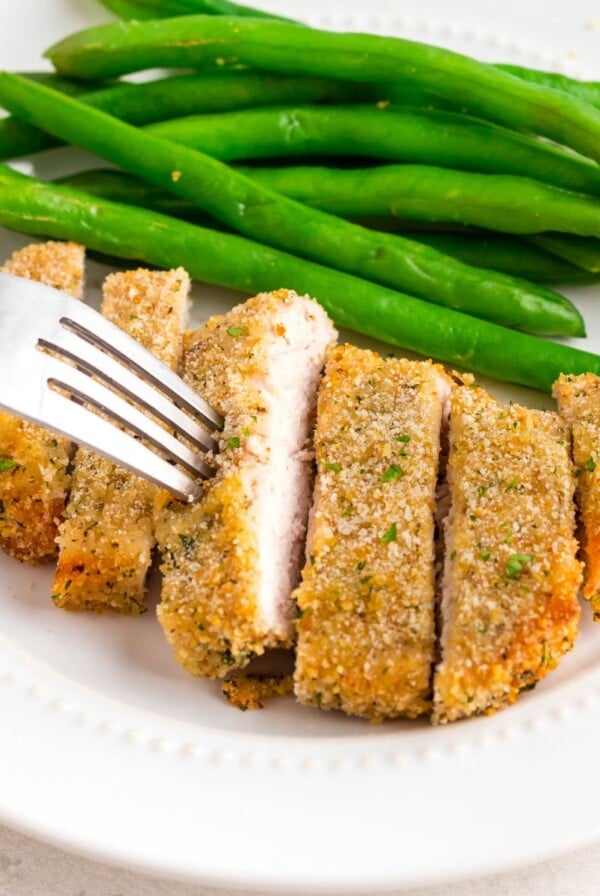 Golden breaded pork chop on a white plate with green beans as a side.