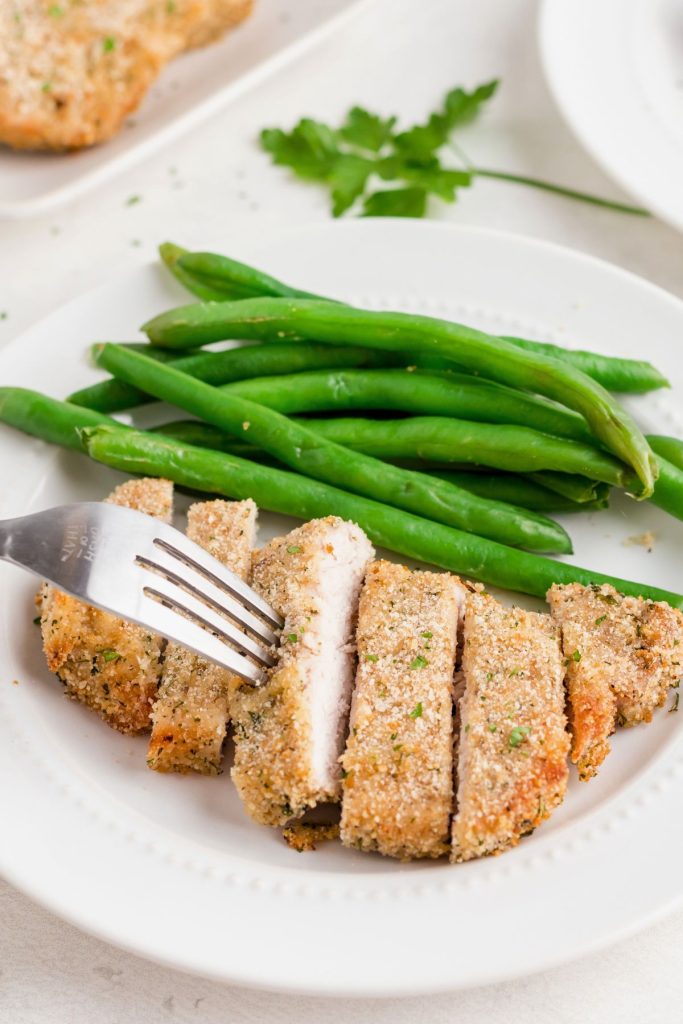 Breaded pork chop on a white plate, cut into slices and served with green beans. 