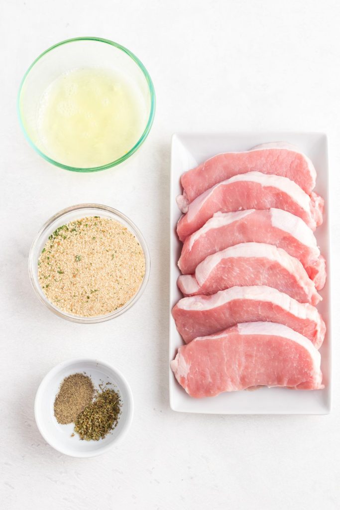 Ingredients needed to make breaded pork chops on a white marble table. 