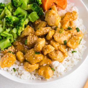 Juicy orange chicken served over rice with broccoli on a white plate.