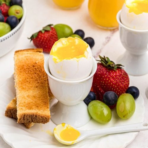 Slightly runny yolk inside of an egg shell in an egg holder with fruit and toast.