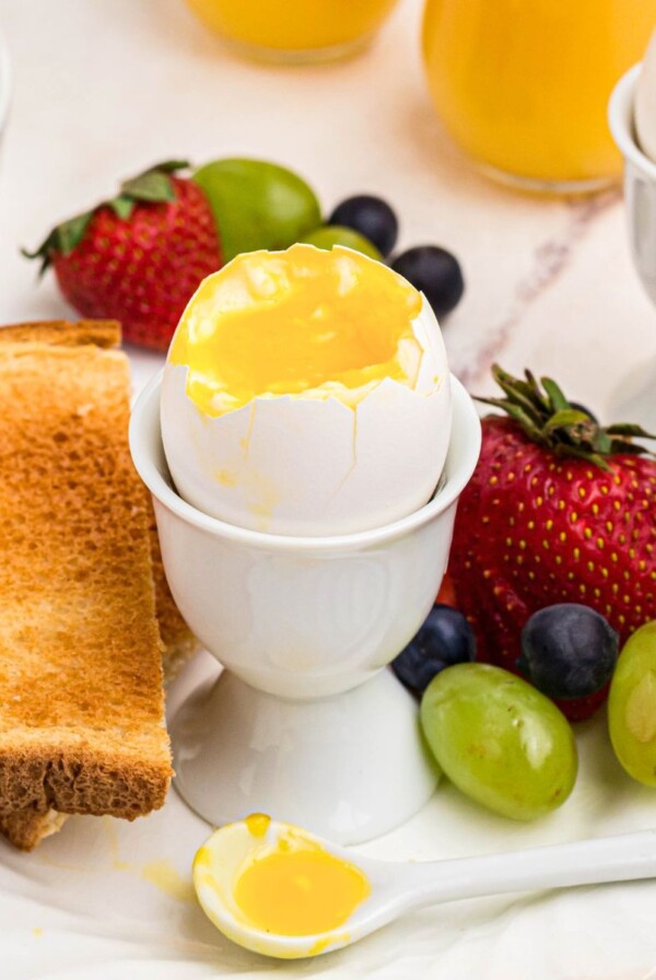 Slightly runny yolk inside of an egg shell in an egg holder with fruit and toast.