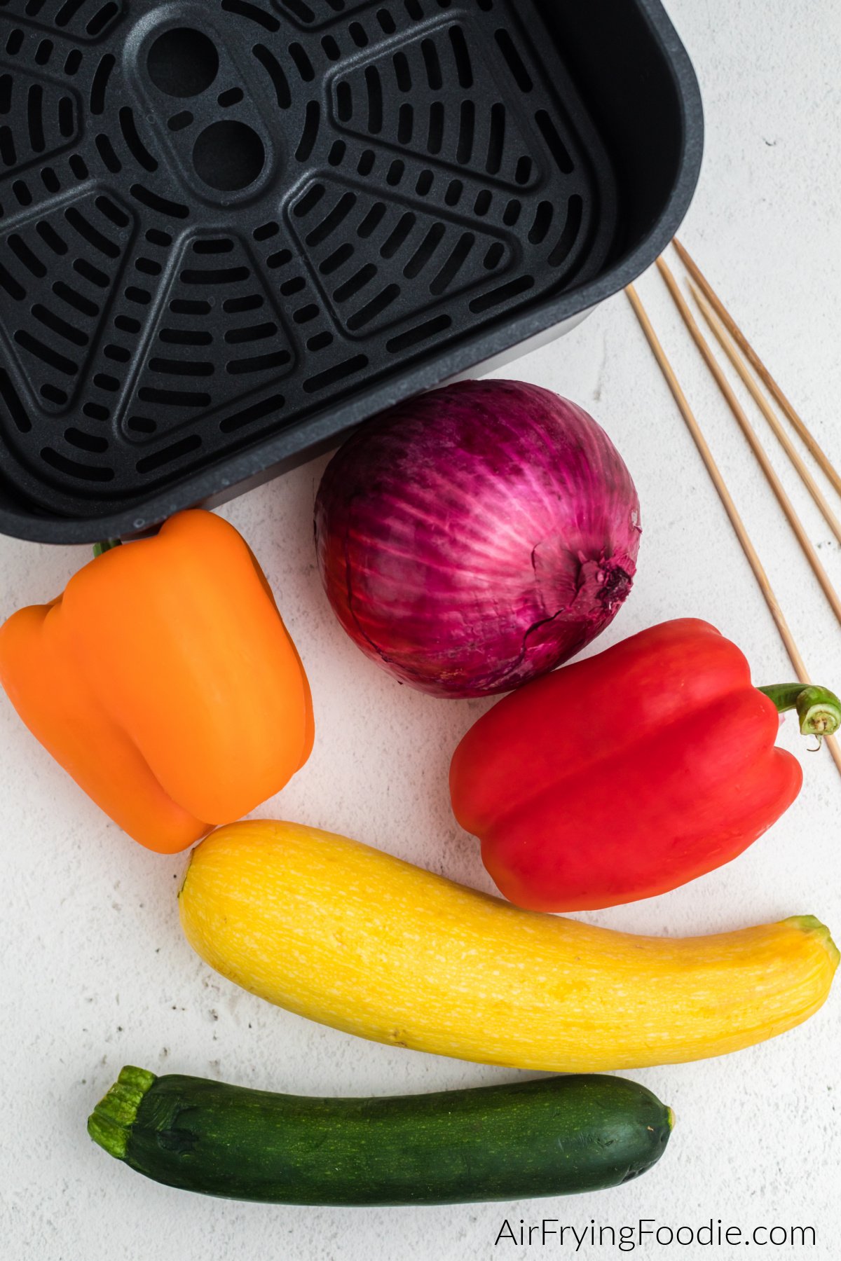 Orange and red bell pepper, red onion, yellow squash, and zucchini squash with skewers and an air fryer basket.