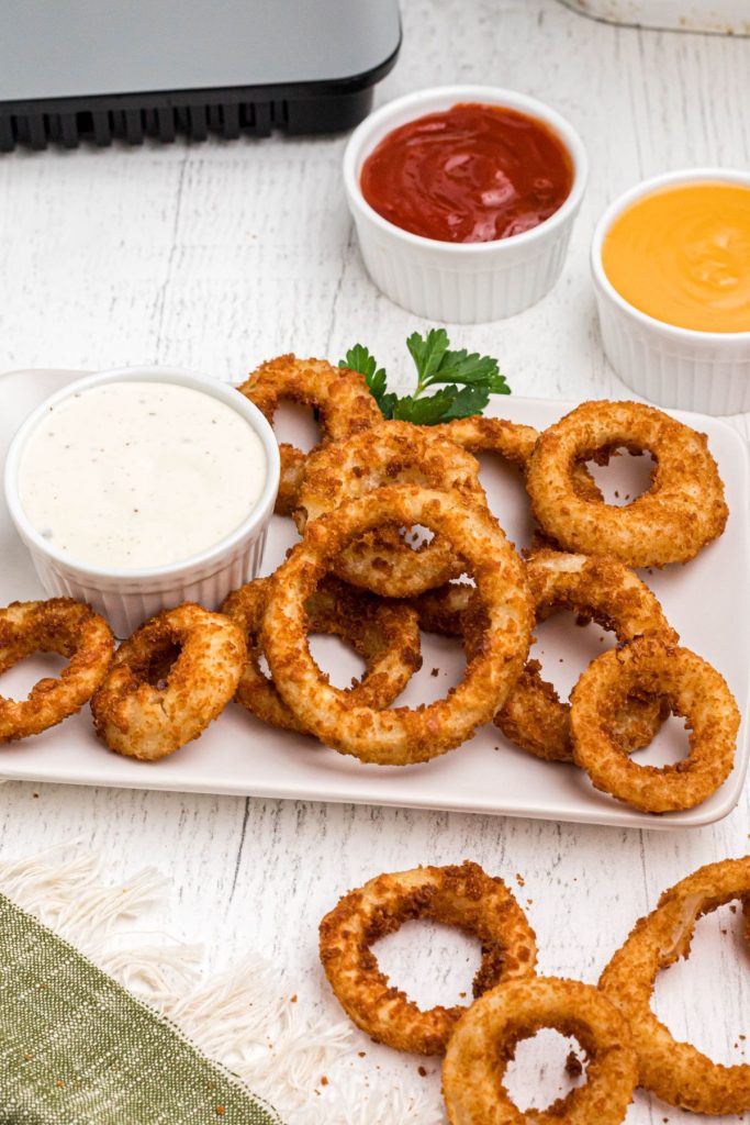 Golden crispy air fried onion rings stacked on a white plate served with ranch and other sauces. 