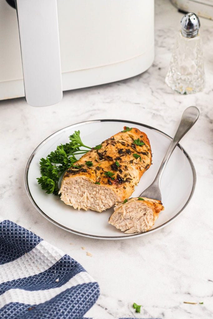 One chicken breast on a small white plate, garnished with parsley in front of a white air fryer. 