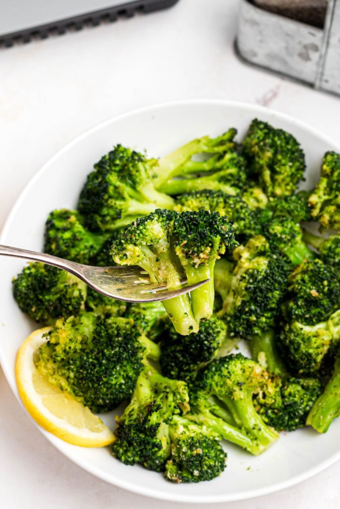 Juicy broccoli on a fork over a plate of broccoli with a lemon wedge. 