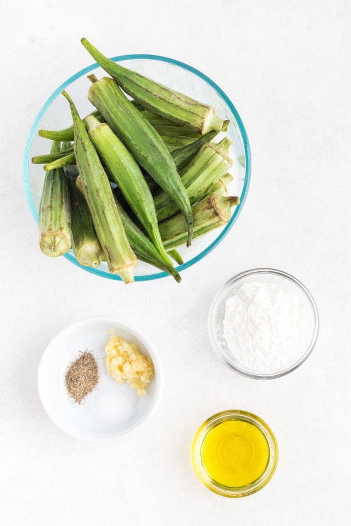Uncooked okra, olive oil, garlic, seasonings, and cornstarch, measured in bowls on a white marble table. 