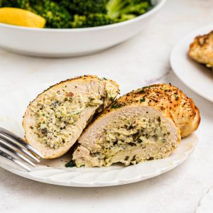 Golden stuffed chicken on a white plate with a bowl of broccoli behind the plate.