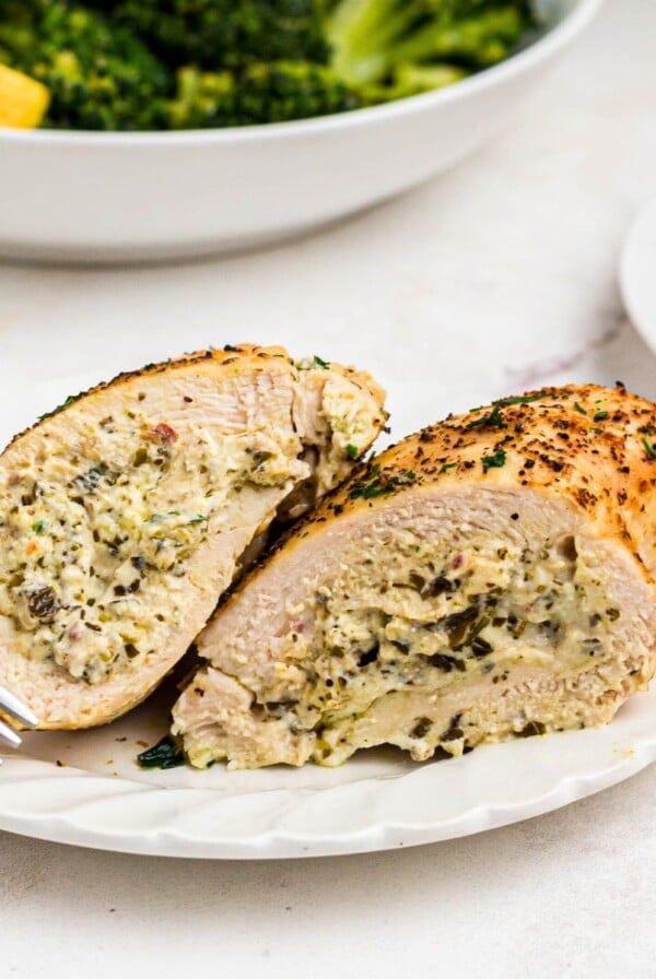 Golden stuffed chicken on a white plate with a bowl of broccoli behind the plate.