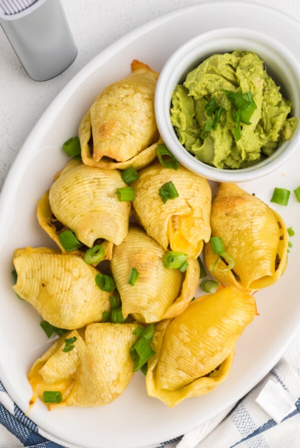 Taco stuffed shells on a plate topped with green onions and served with a side of guacamole.