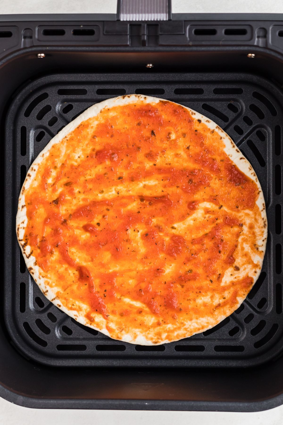 Tortilla in the air fryer basket with pizza sauce spread on top.