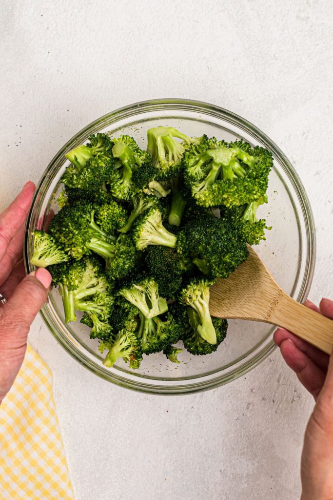 Uncooked broccoli in a large clear glass bowl being tossed with olive oil and seasonings. 