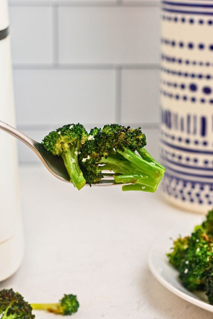 Green seasoned broccoli florets on a fork in front of an air fryer. 