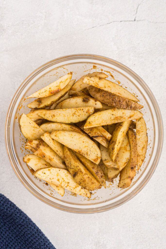 Uncooked potato wedges seasoned in a clear glass bowl.