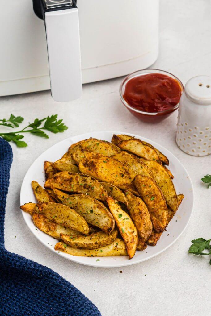 Golden seasoned potato wedges on a white plate in front of a white air fryer. 