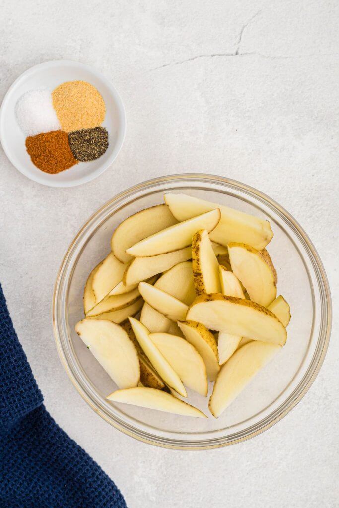 Uncooked potato wedges in a clear glass bowl with seasonings in a small dish on a marble table. 