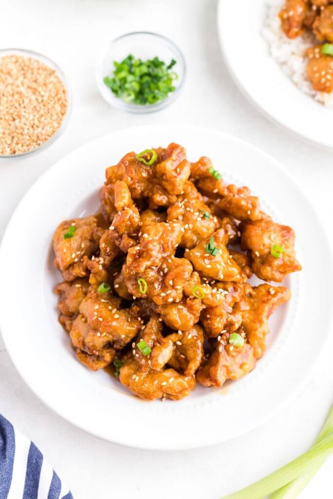 Golden orange chicken pieces in a white bowl topped with sesame seeds and chopped green onion. 