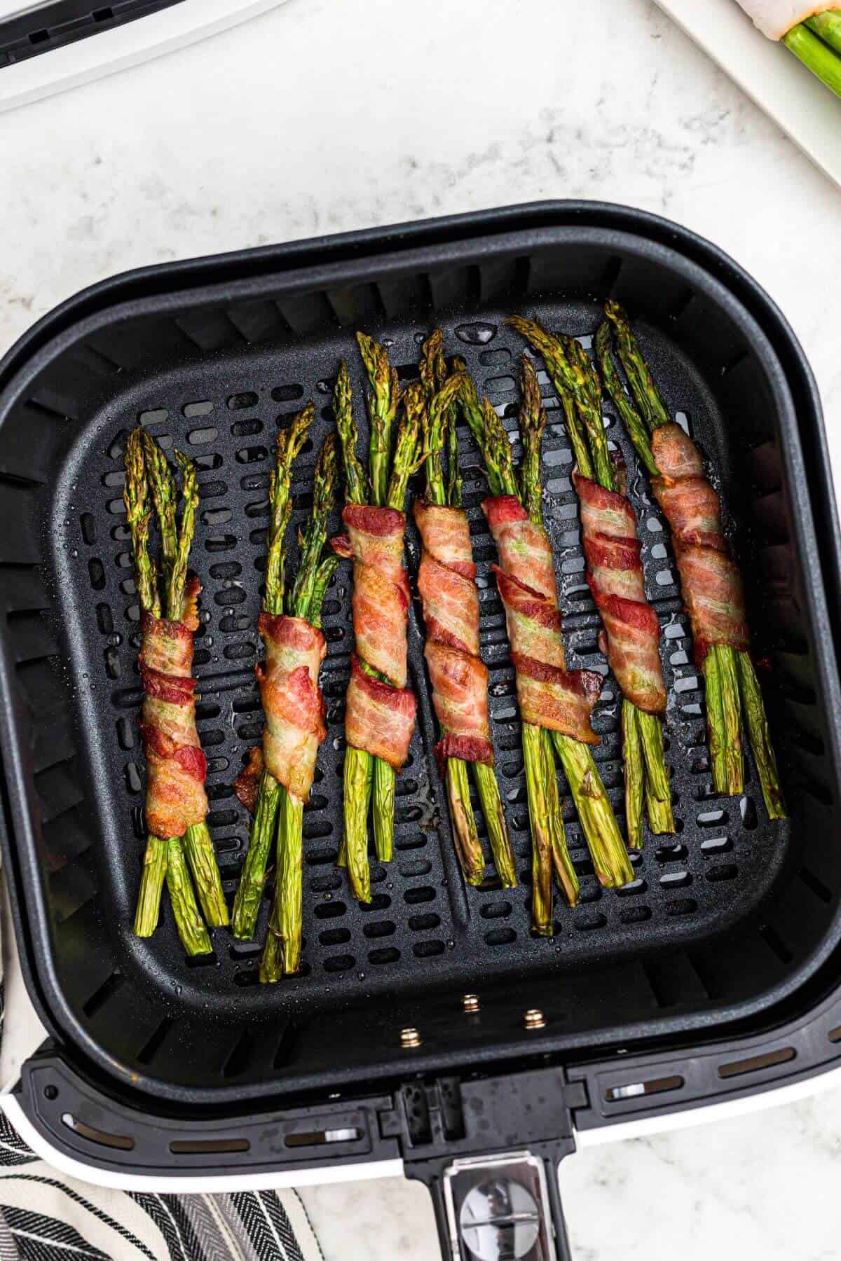 Crispy brown bacon strips wrapped around asparagus stalks in the air fryer basket after being cooked. 